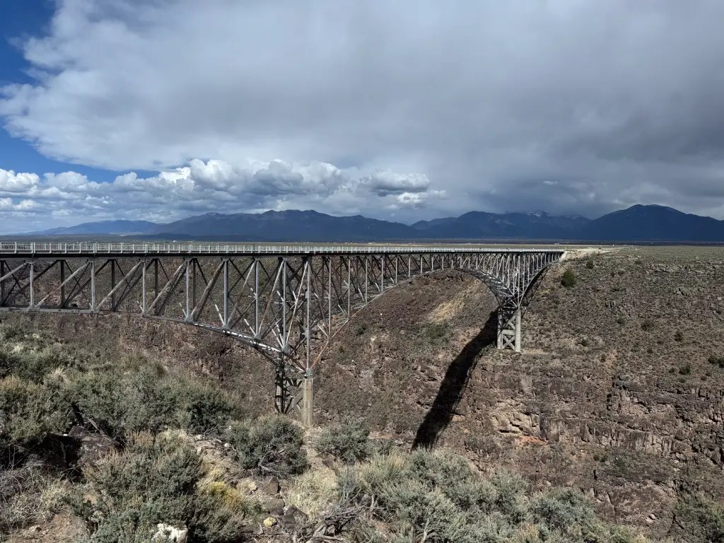 A wide view of the Rio Grande Gorge Bridge, showcasing its impressive steel structure spanning a deep canyon, with mountains and dramatic clouds in the background.