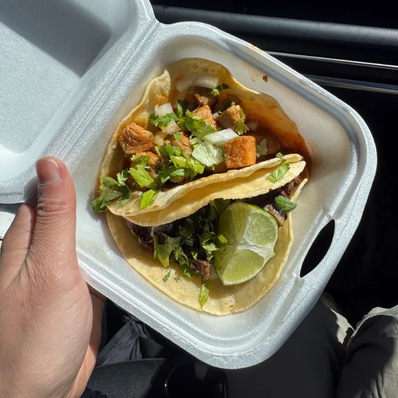 A hand holding a takeout container with two tacos topped with diced meat, onions, and cilantro, accompanied by a lime wedge.