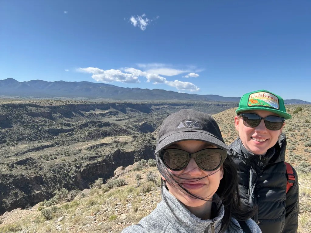 A scenic view of the Rio Grande Gorge in New Mexico, featuring two people in the foreground wearing sunglasses and hats. They are enjoying the outdoors with mountains and blue skies in the background.