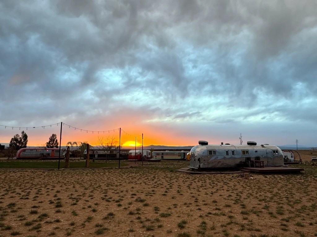 A scenic view of vintage Airstream trailers at sunset, surrounded by open desert landscape and dramatic, colorful clouds in the sky.