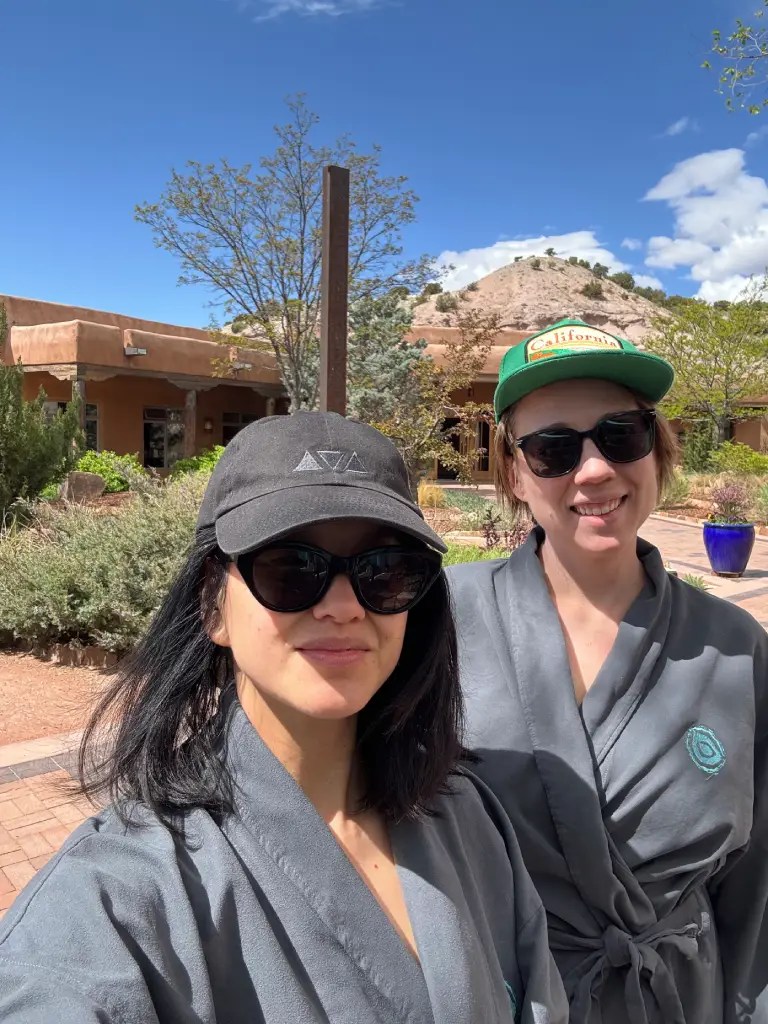 Two women in bathrobes posing for a selfie outdoors at a spa resort, surrounded by greenery and a blue sky.