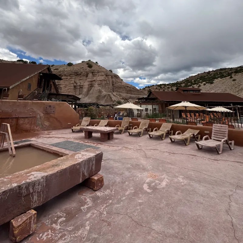 Outdoor spa area at Ojo Caliente Mineral Springs Resort featuring lounge chairs, a mud bath, and sandstone cliffs in the background under a partly cloudy sky.