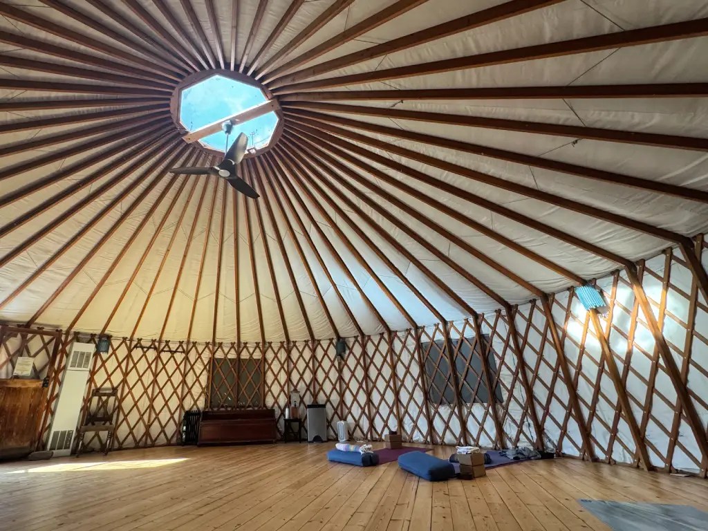 Interior view of a yurt with a wooden ceiling, featuring beams radiating from a central skylight and yoga mats arranged on the floor.