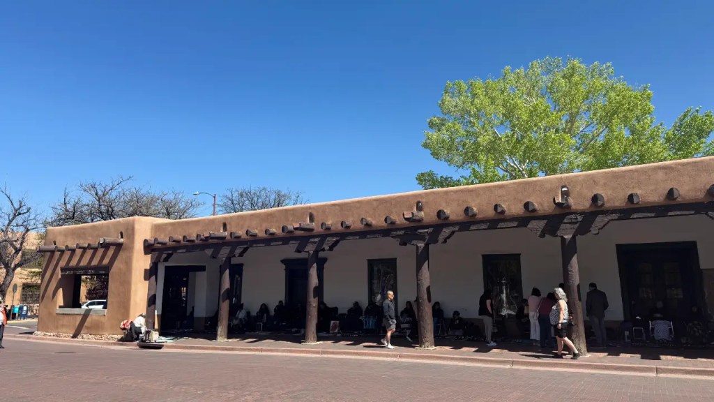 A historic adobe building in Santa Fe, New Mexico, featuring a wooden beam porch, people gathering outside, and a clear blue sky.