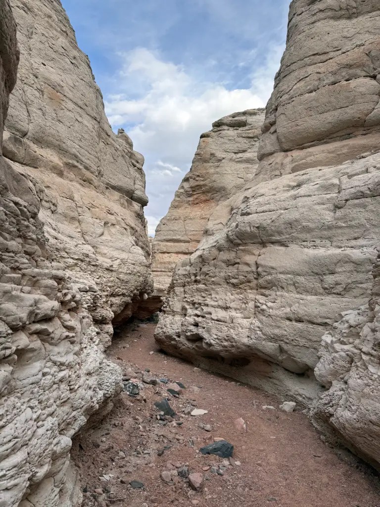Narrow canyon pathway with towering cream-colored sandtone rock formations on either side, under a cloudy blue sky.