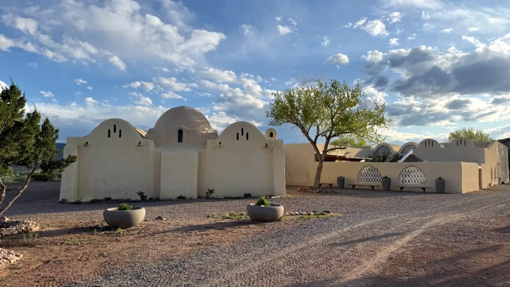 Exterior view of a white adobe-style mosque with rounded roofs and decorative elements, surrounded by gravel and greenery under a partly cloudy sky.
