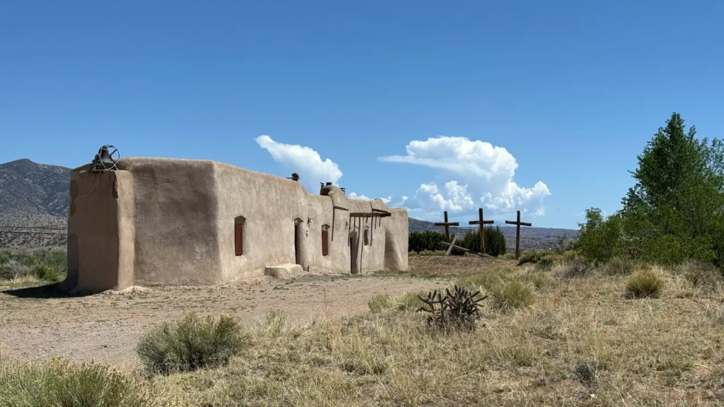 Adobe building with wooden window shutters and crosses in the background, set against a clear blue sky and desert landscape.