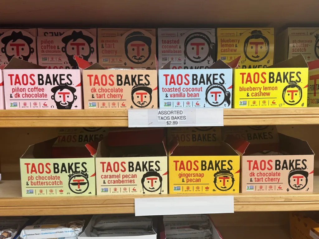Display of Taos Bakes snack boxes featuring various flavors including piñon coffee, dark chocolate, blueberry lemon, and caramel pecan on a wooden shelf.