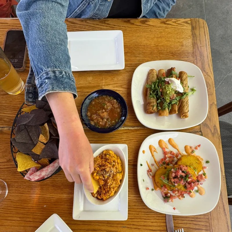A person reaching for a plate of food with a fork, surrounded by various dishes including rolled tacos, a bowl of salsa, and a side of nachos, all set on a wooden table.