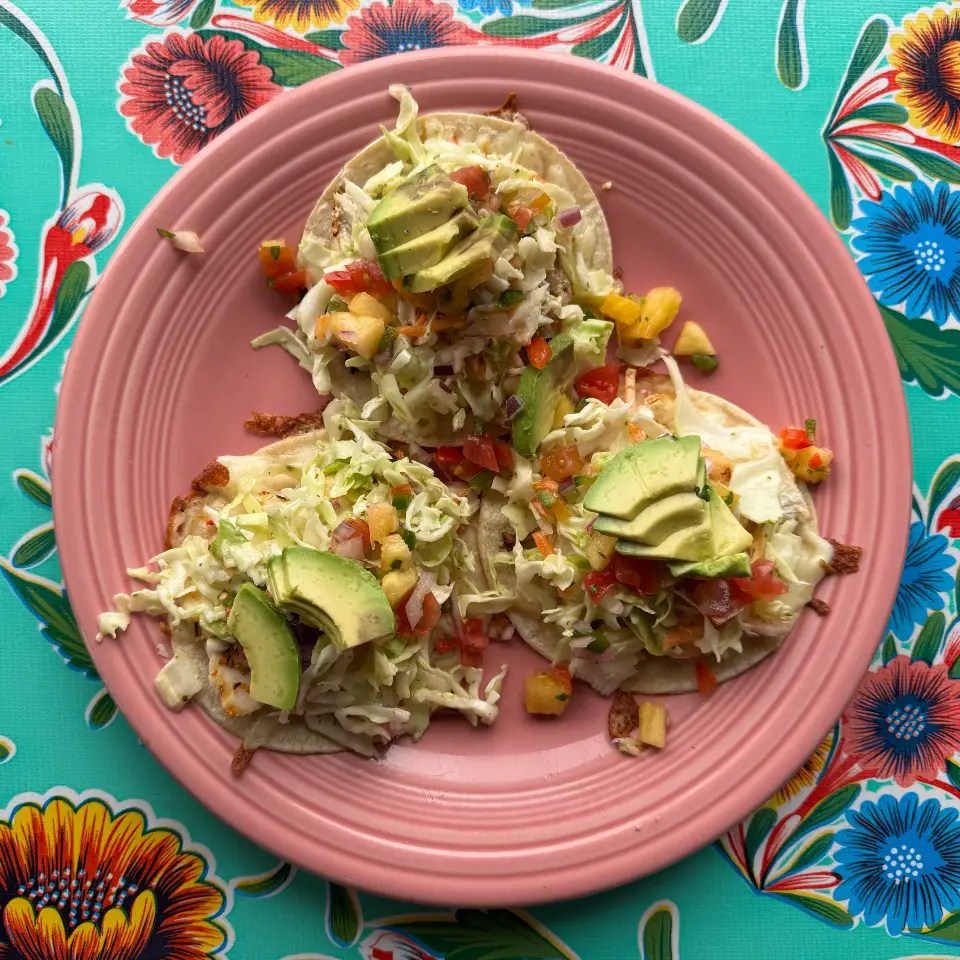 A plate of three fish tacos topped with shredded cabbage, diced tomatoes, and slices of avocado, served on a pink plate against a colorful floral background.