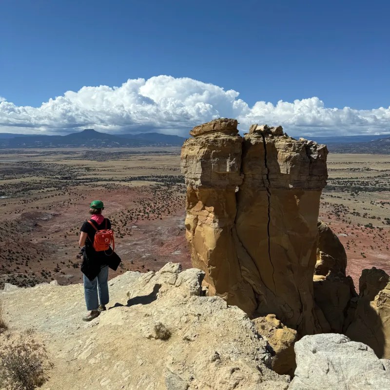 A woman standing on a rocky ledge, overlooking Chimney Rock  and desert terrain under a bright blue sky with fluffy white clouds.