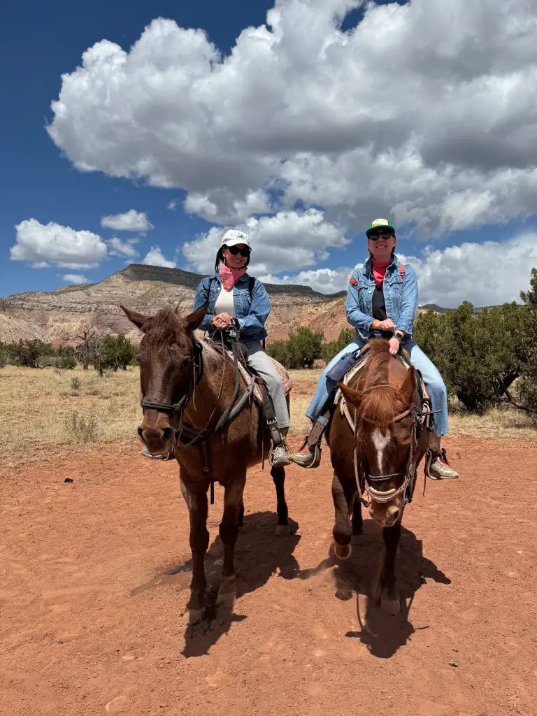 Two women riding horses in a dusty landscape with scattered bushes and a backdrop of mountains under a blue sky with white clouds.