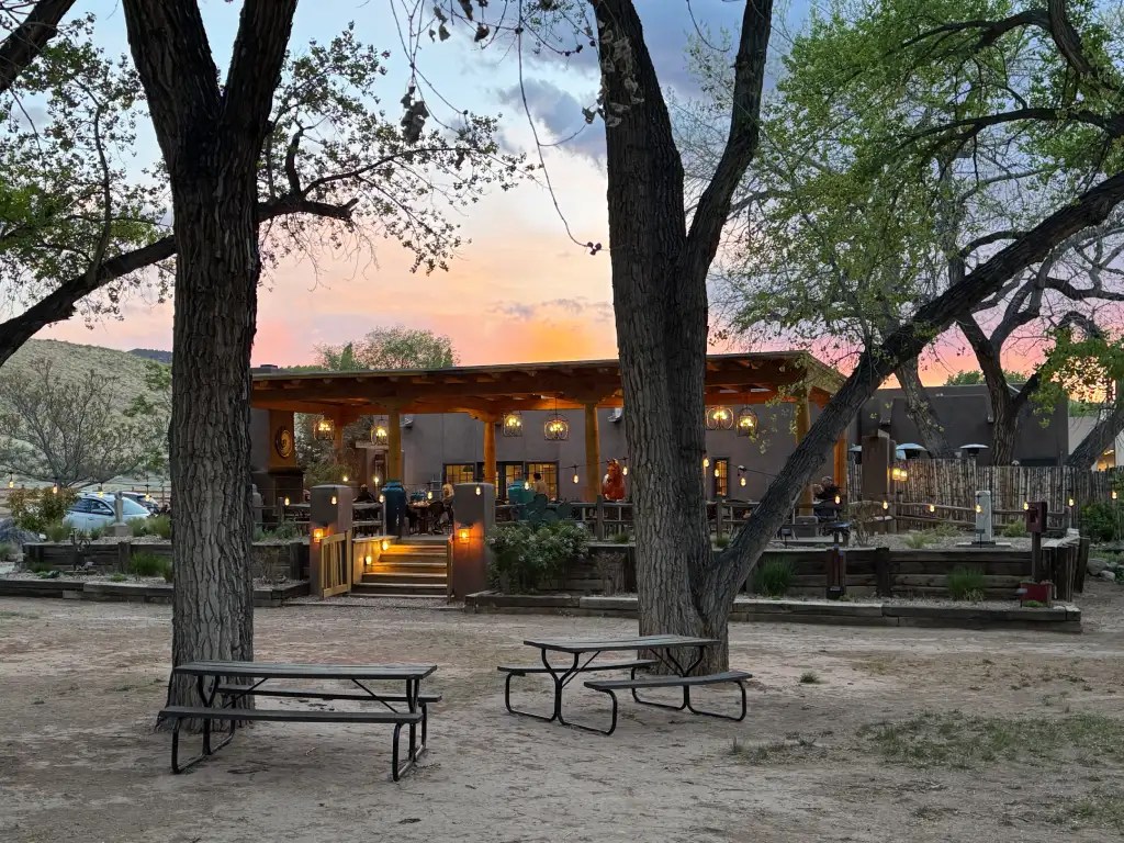 Exterior view of a rustic restaurant with a patio, surrounded by trees and a sunset sky, featuring picnic tables in the foreground.