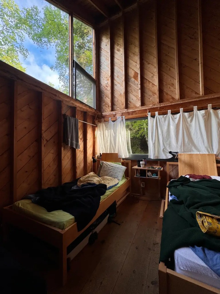 Interior view of a rustic dorm room at Haystack, featuring wooden walls, two twin beds with bedding, a bedside table, and large windows providing natural light.