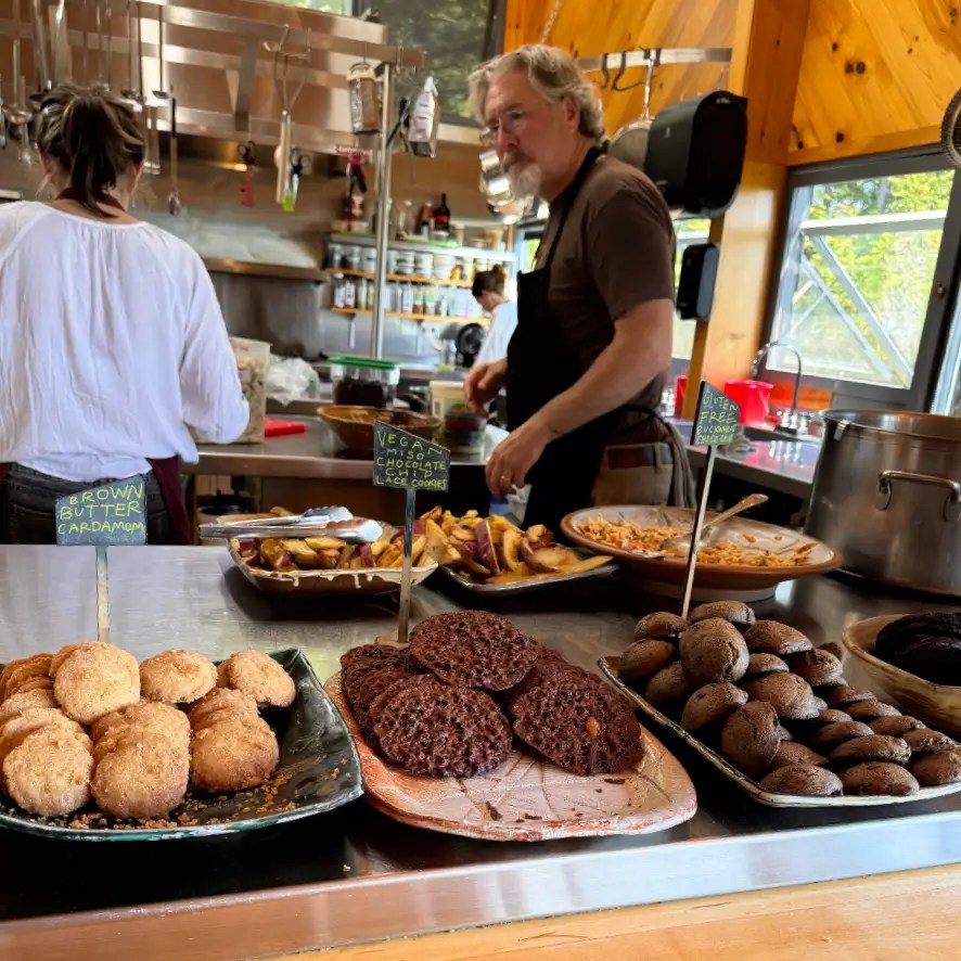 A peek into the kitchen at Haystack School, featuring an assortment of homemade cookies, including brown butter cardamom cookies and vegan chocolate chip lace cookies, with a chef working in the background.