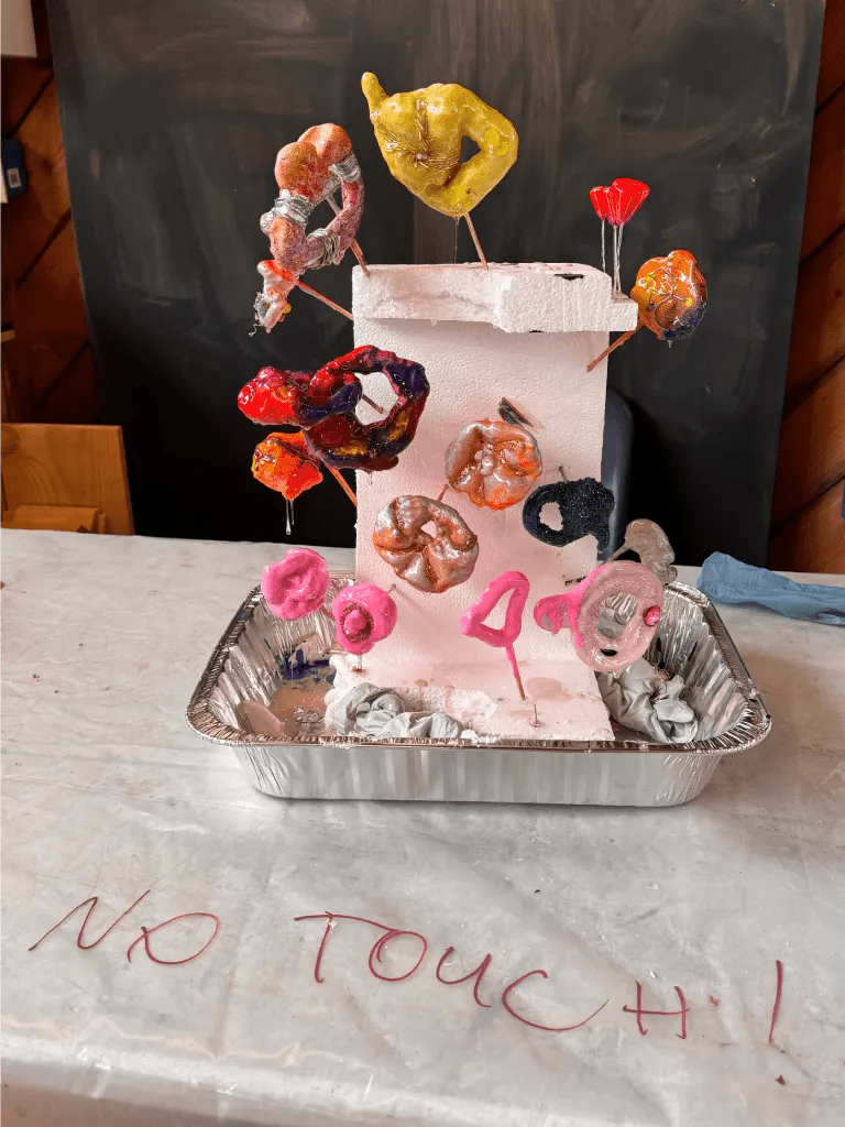 An artistic display featuring colorful jewelry pieces made from resin and other materials, mounted on a foam structure, with the words 'NO TOUCH!' written in pink on the tablecloth underneath.