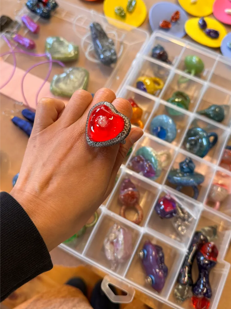 A person wearing a red heart-shaped ring, surrounded by a variety of colorful jewelry pieces in a compartmentalized tray.