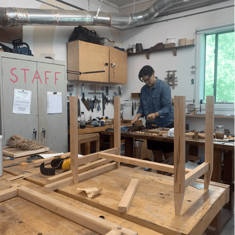 A woodworking studio with a partially assembled wooden bench in the foreground and a person working on a project in the background, surrounded by various tools and materials.