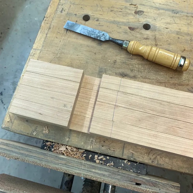 A close-up view of woodworking tools and materials on a workbench, featuring a chisel and wooden pieces with marked lines for cutting.