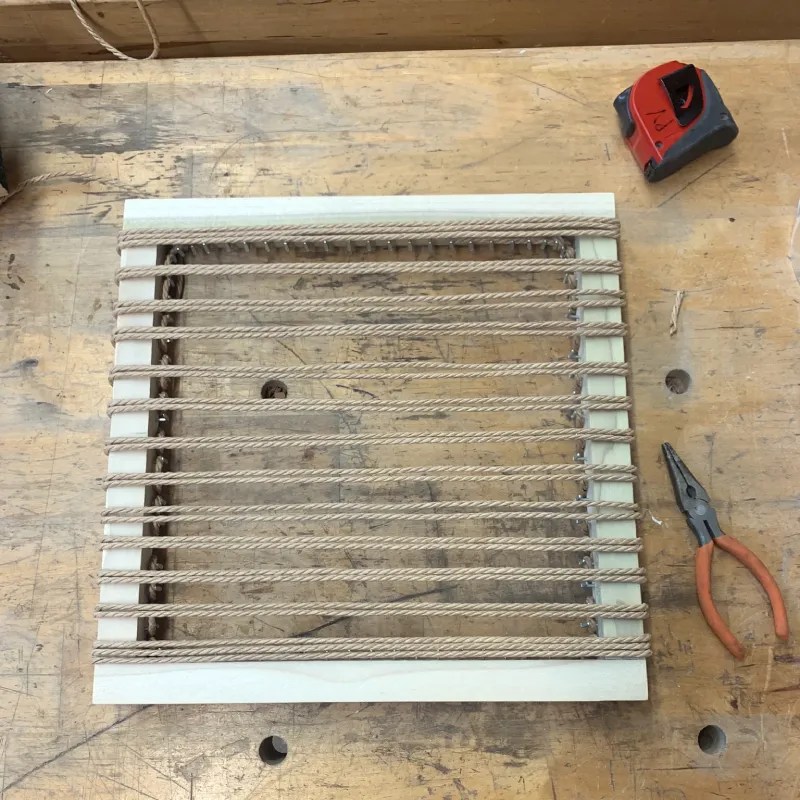 A wooden frame with woven Danish cord on a workbench, accompanied by a pair of pliers and a measuring tape.