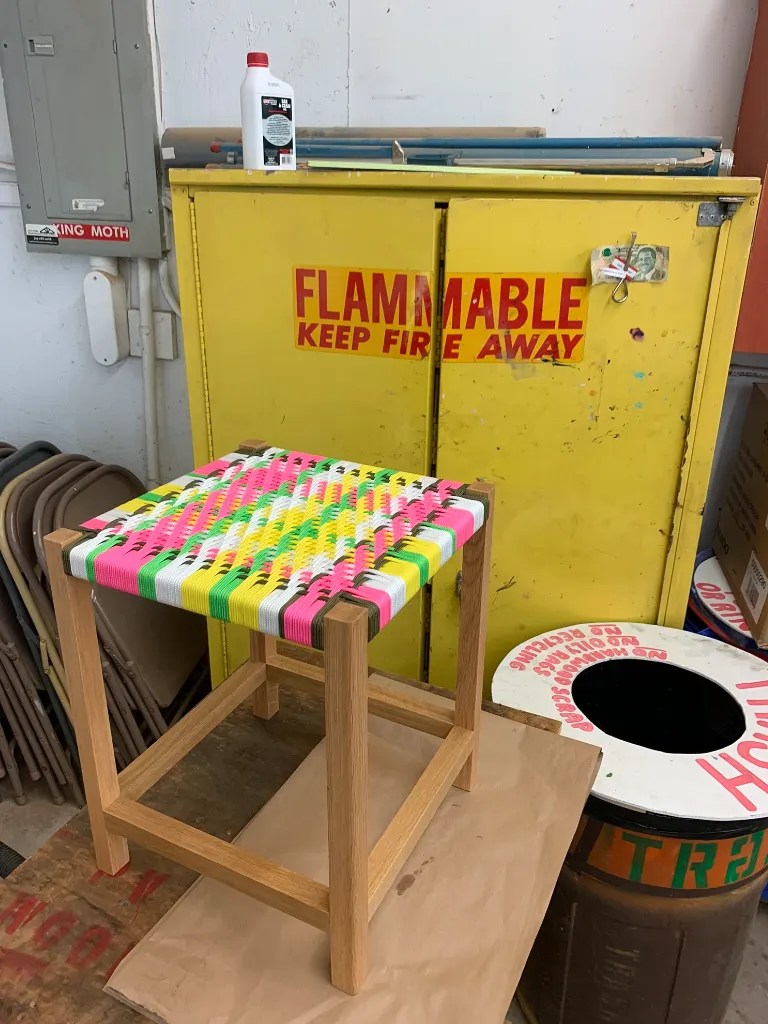 A colorful woven stool made of wood and polypropylene cord, sitting in a workshop near a bright yellow storage cabinet stamped with the word "flammable".