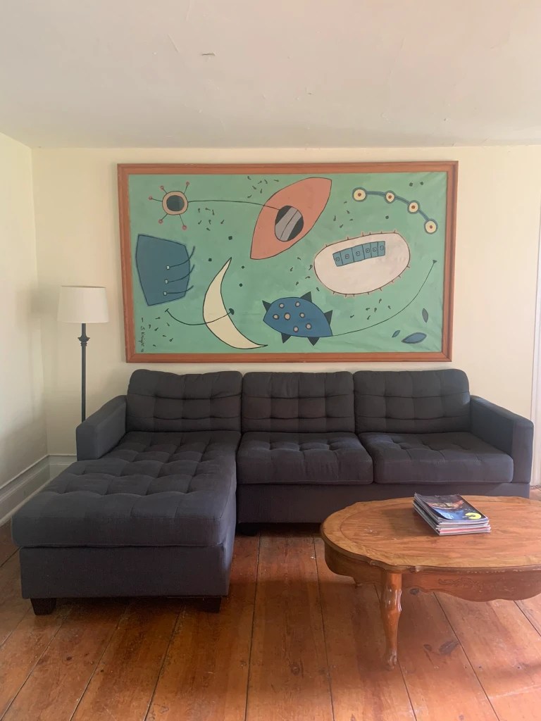 A cozy living room featuring a dark gray sectional sofa, a wooden coffee table with magazines, and a large abstract painting on the wall with colorful shapes against a light green background.