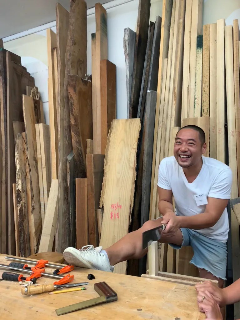 A man smiling and demonstrating woodworking techniques in a studio filled with various pieces of wood, with tools on a workbench in front of him.