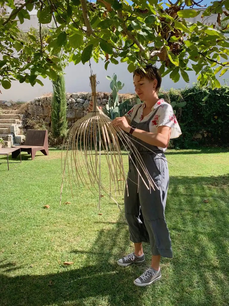 A woman weaving a lampshade from natural materials in a sunny outdoor setting, surrounded by greenery and the shadows of trees.