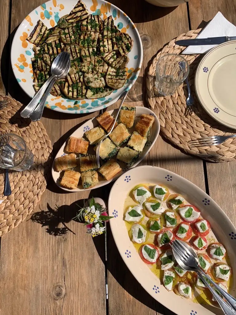 A beautifully arranged outdoor meal featuring grilled zucchini, flaky pastries, and a platter of mozzarella wrapped in basil leaves, set on a rustic wooden table.