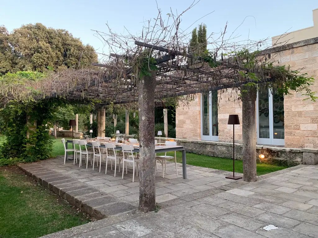 A serene outdoor dining area at a villa in Italy, featuring a long table set for a meal under a vine-covered pergola, surrounded by lush greenery.