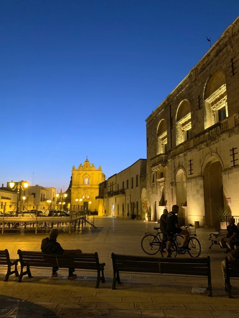 A picturesque evening view of a quiet square in Muro Leccese, Italy, with illuminated historic buildings and a few people sitting on benches.