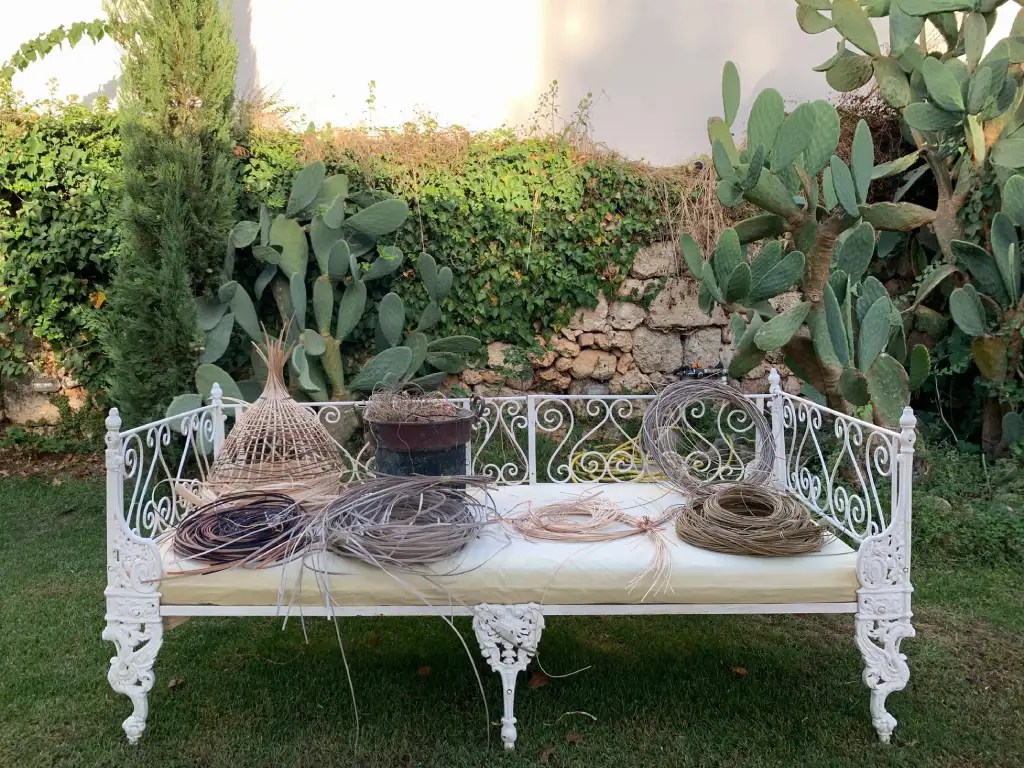 A beautifully decorated outdoor bench with various weaving materials arranged on it, surrounded by a lush garden with cactus plants in the background.