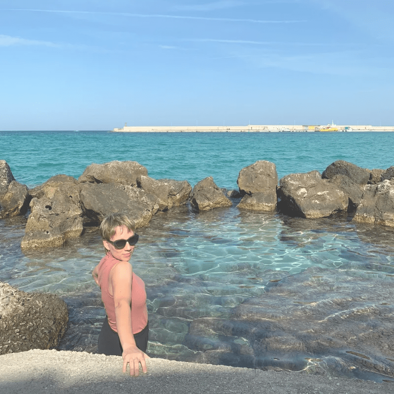 A woman standing in clear blue water near a rocky shoreline, wearing sunglasses and a sleeveless top, with a harbor visible in the background.