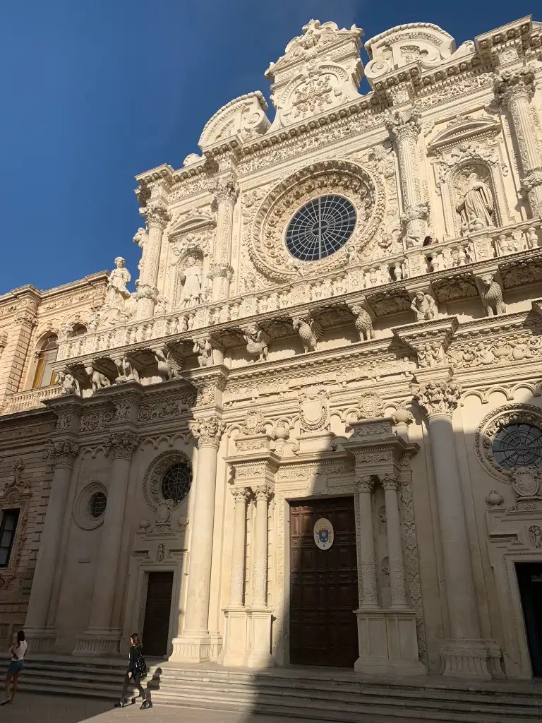 Facade of the Basilica of the Holy Cross in Lecce with intricate sculptures and carvings, featuring a prominent circular window and tall wooden door, set against a clear blue sky.
