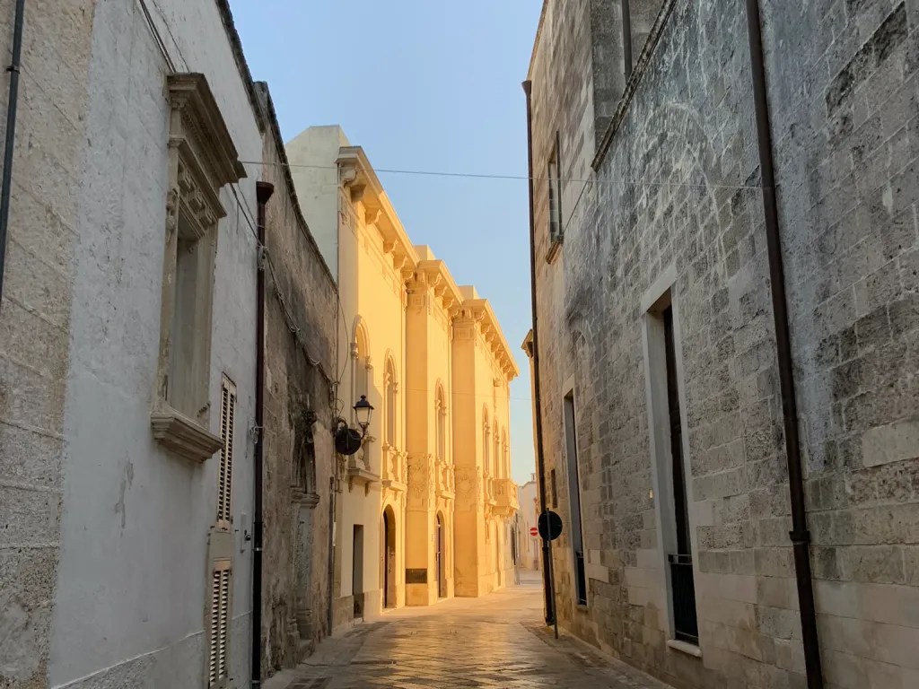 Narrow cobblestone street in Muro Leccese, Italy, with sunlight illuminating a historic building at the end.