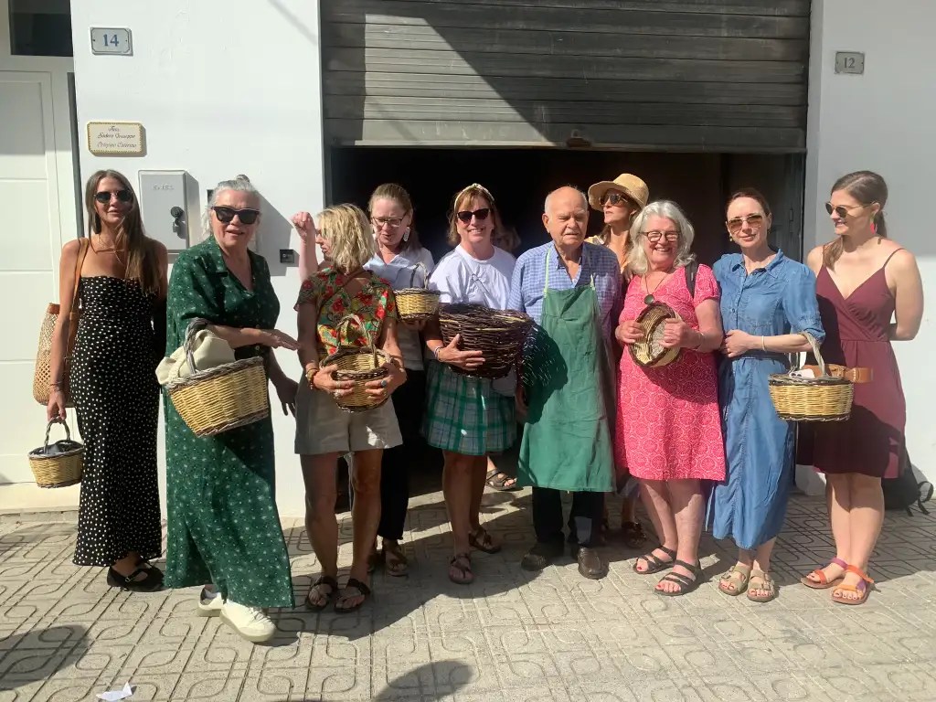 A group of women, including a man in an apron, standing outside with woven baskets in their hands, smiling together in a sunny setting.