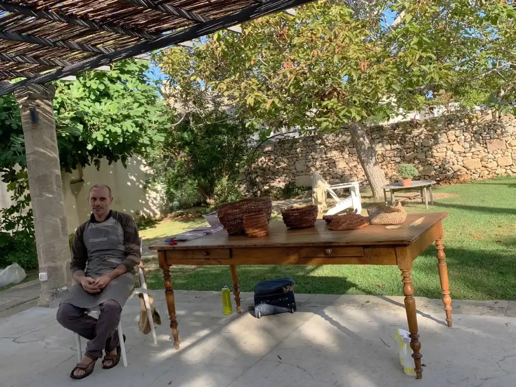 An instructor seated at a table demonstrating basket weaving techniques outdoors, surrounded by greenery and woven baskets.