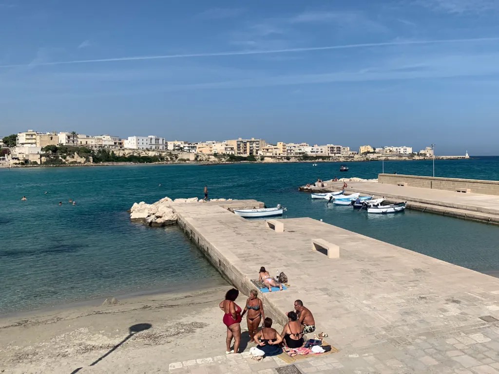 A coastal view featuring a sandy beach with several people relaxing and swimming in the water, a stone pier extending into the sea, and small boats docked nearby, with a backdrop of buildings along the shore under a blue sky.