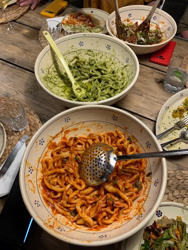 A beautifully arranged table with bowls of colorful Italian pasta dishes, including green pesto pasta and red tomato sauce pasta, surrounded by salad and drinks.