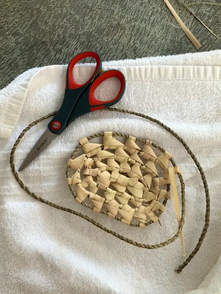 A close-up of scissors and woven palm leaf craft materials placed on a white towel, featuring a partially completed woven basket and a length of twine.