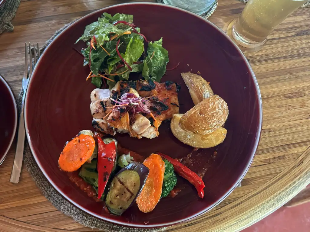Grilled chicken with sautéed vegetables and a fresh salad, served on a deep red plate, accompanied by a cold drink.