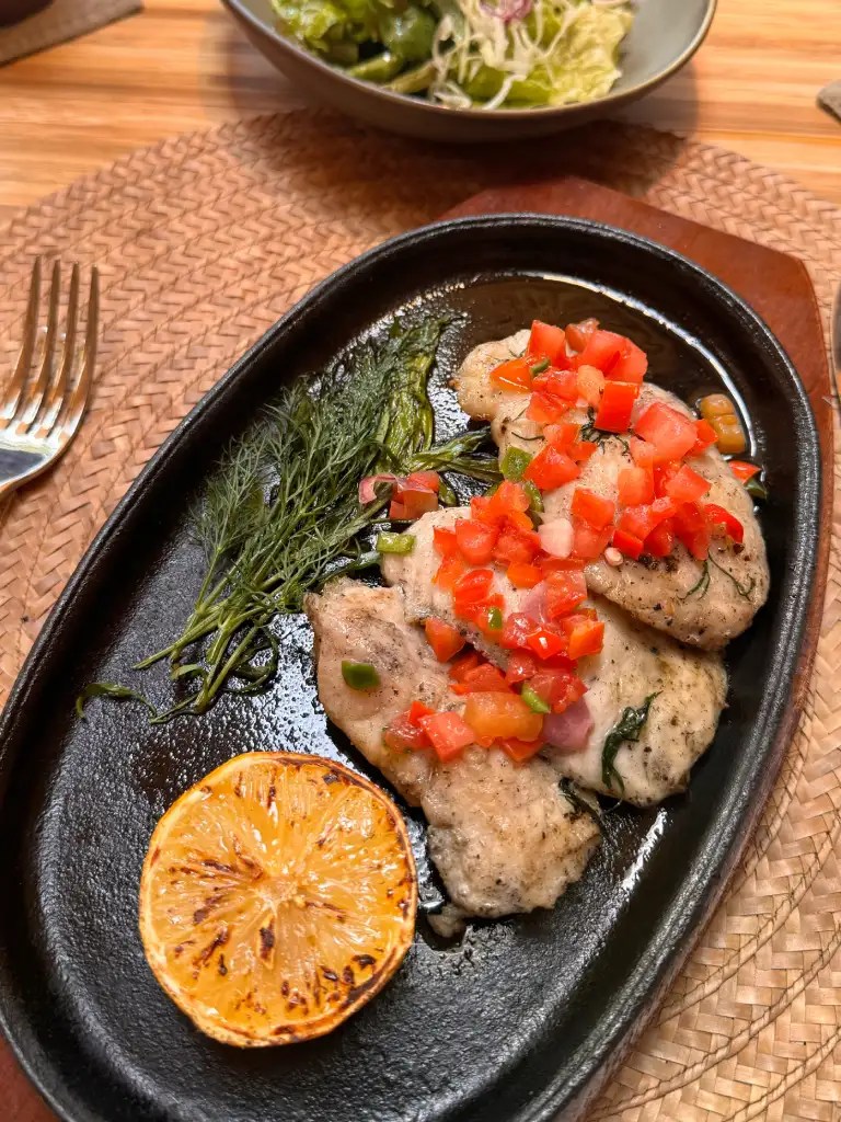 A beautifully presented plate of grilled fish topped with fresh diced tomatoes and herbs, served on a sizzling hot stone plate alongside a grilled lemon slice and a side salad.