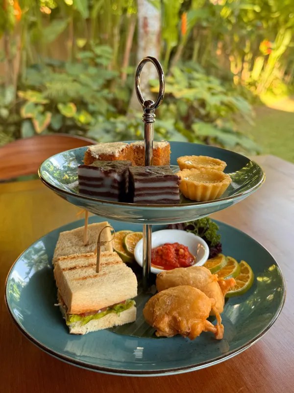 A two-tiered serving platter featuring various traditional snacks and desserts, including sandwiches, fritters, brownies, and small tarts, set against a lush green background.