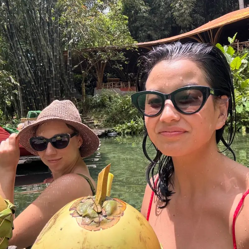 Two women enjoying a sunny day by the water, one holding a coconut, with lush greenery in the background.