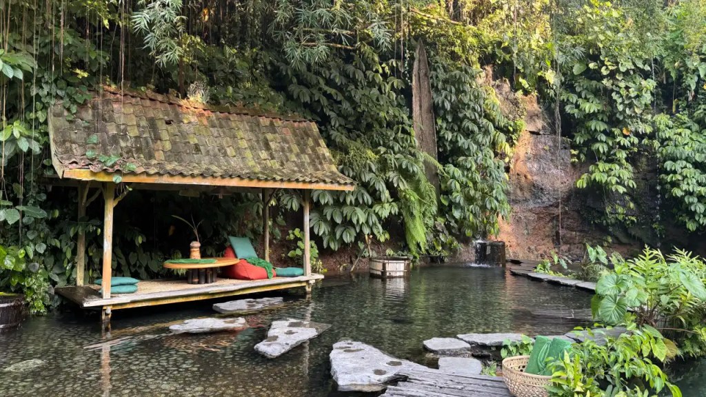 A wooden platform with a thatched roof, surrounded by lush greenery and a calm water pool, featuring colorful cushions for seating.
