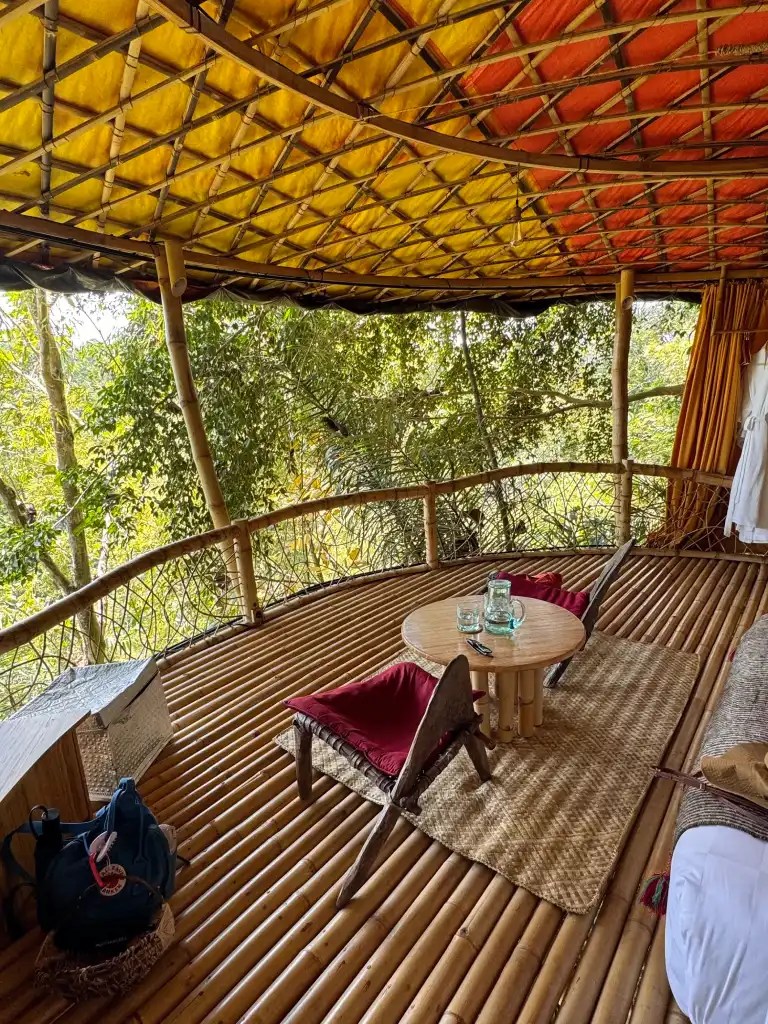 Open-air seating area in a bamboo structure with a colorful rattan roof, surrounded by lush greenery, featuring a round table and cushioned chairs.