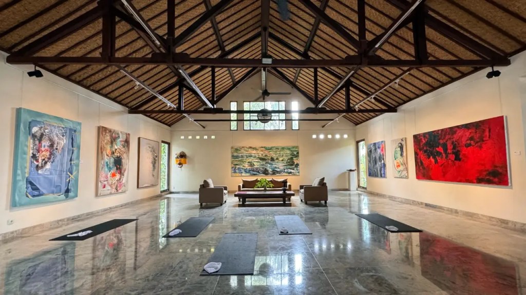 Interior view of an art gallery set up as a yoga studio featuring high ceilings, wooden beams, and marble flooring. Several mats are laid out on the floor, surrounded by colorful artwork on the walls.
