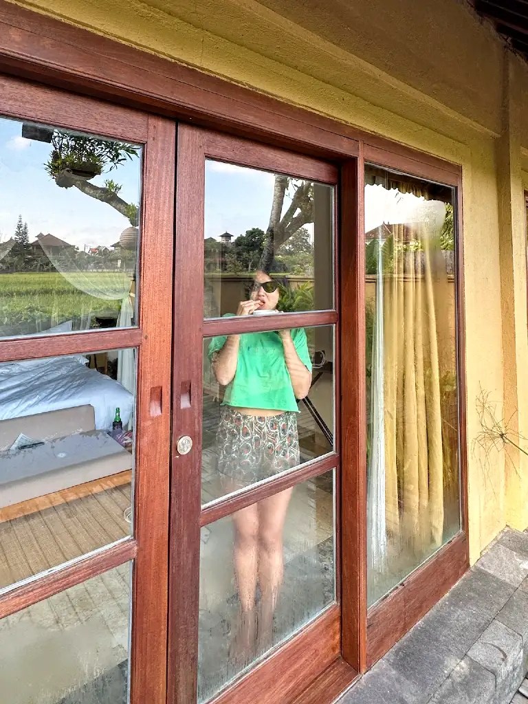 A person standing inside a villa, framed by wooden sliding doors, holding a snack and wearing sunglasses. The background features a glimpse of the room's interior and a view of lush greenery outside.