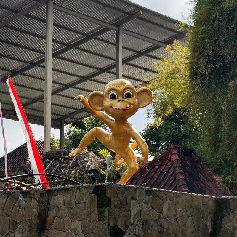 A vibrant, oversized statue of a monkey with a joyful expression, positioned on a stone wall with a thatched roof structure in the background.