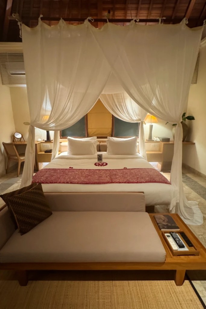 Interior view of a cozy hotel room featuring a king-sized bed with mosquito netting, a seating area with cushions, and decorative elements that evoke a serene ambiance.